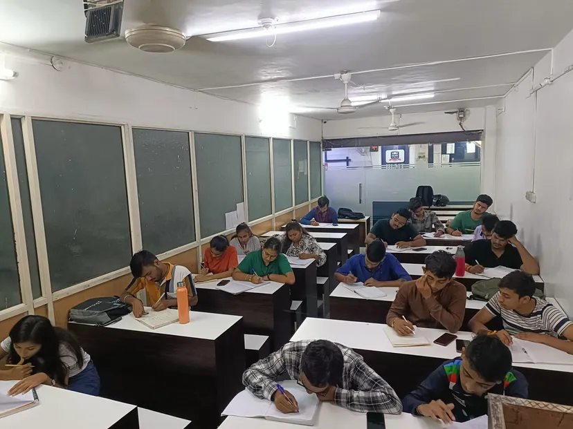 A full classroom mid-session: around fifteen students writing at their desks, green boards along the wall behind them